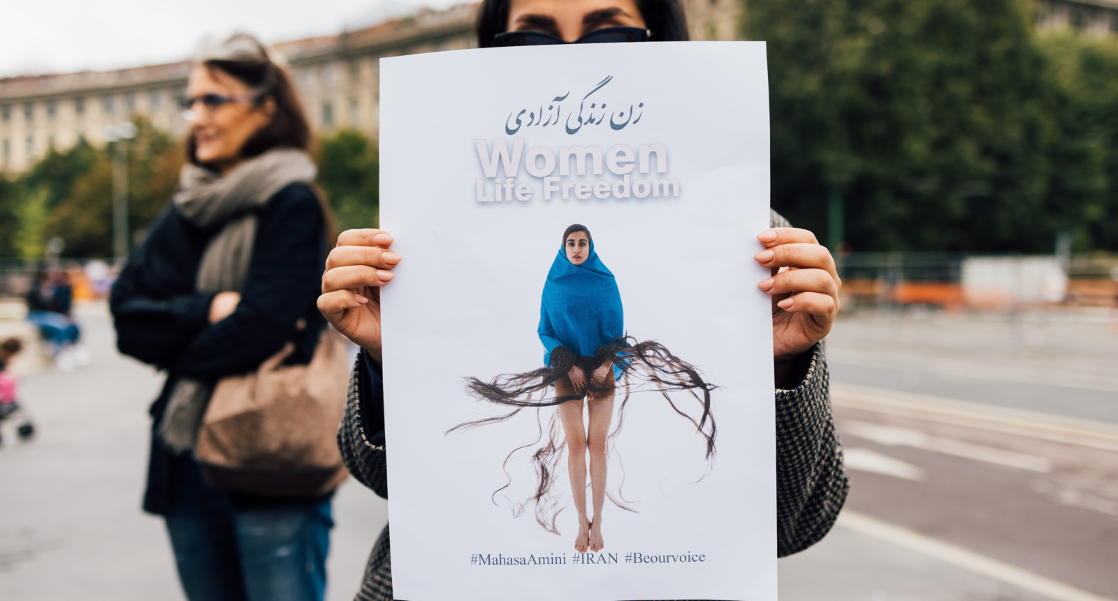 A person stands outdoors holding up a large protest poster in front of their face. The poster features a woman draped in a blue garment with long flowing hair strands extending outward, symbolically. At the top of the poster are the words “Women Life Freedom” in English and Persian. Hashtags at the bottom read #MahsaAmini #IRAN #Beourvoice. Another person is seen standing in the background with arms crossed, and there are buildings and trees in the distance.