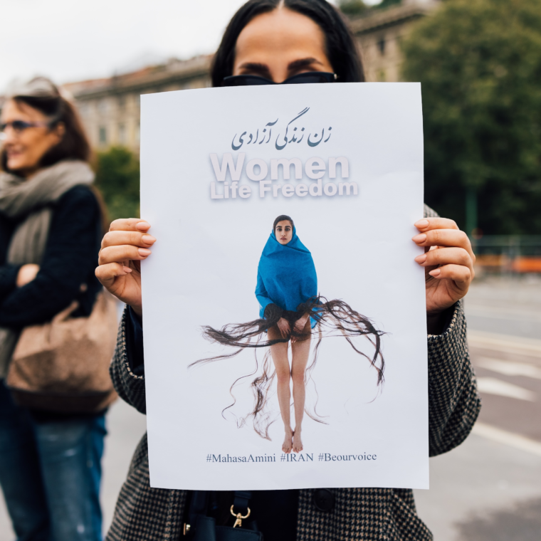 A person stands outdoors holding up a large protest poster in front of their face. The poster features a woman draped in a blue garment with long flowing hair strands extending outward, symbolically. At the top of the poster are the words “Women Life Freedom” in English and Persian. Hashtags at the bottom read #MahsaAmini #IRAN #Beourvoice. Another person is seen standing in the background with arms crossed, and there are buildings and trees in the distance.