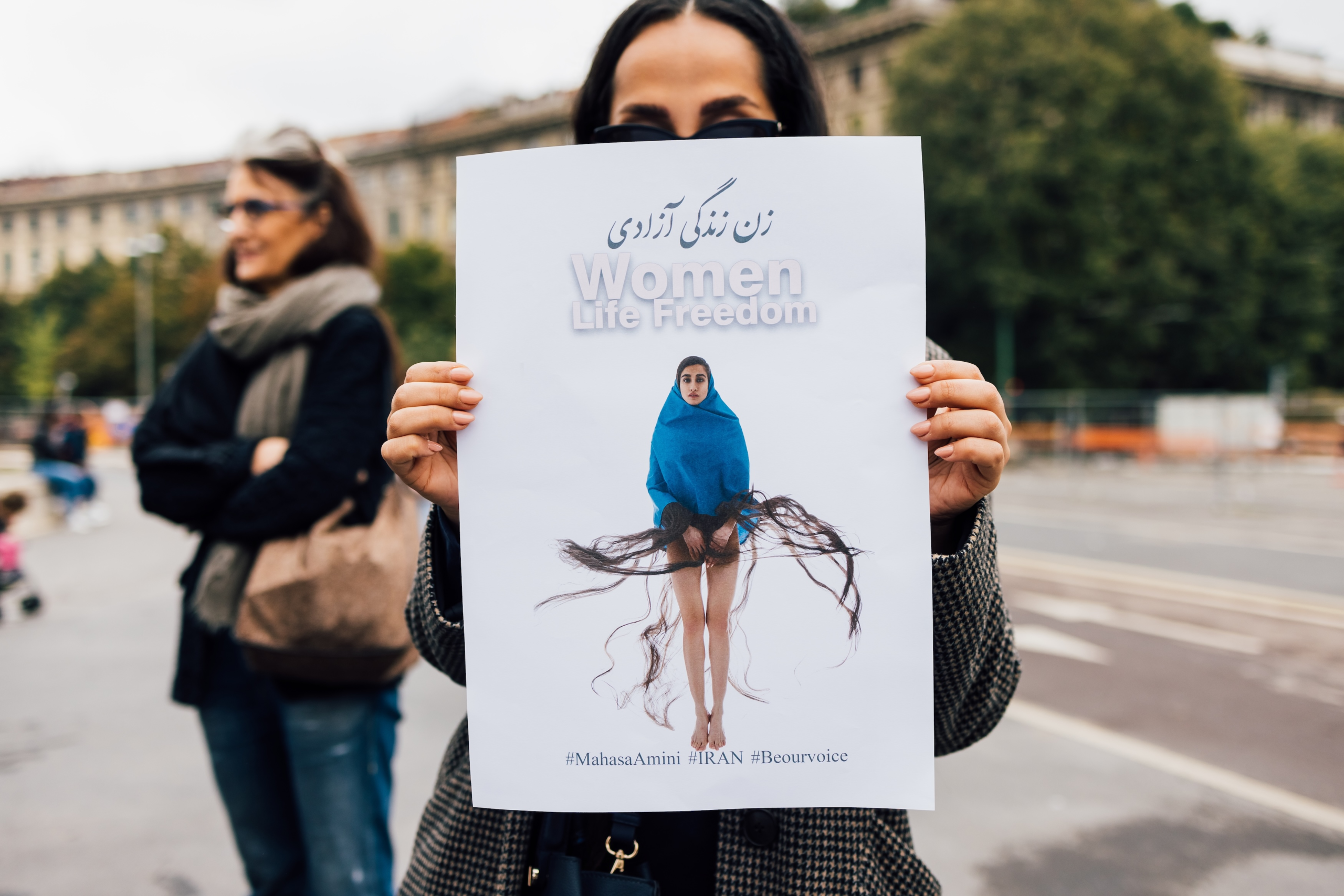 A person stands outdoors holding up a large protest poster in front of their face. The poster features a woman draped in a blue garment with long flowing hair strands extending outward, symbolically. At the top of the poster are the words “Women Life F...