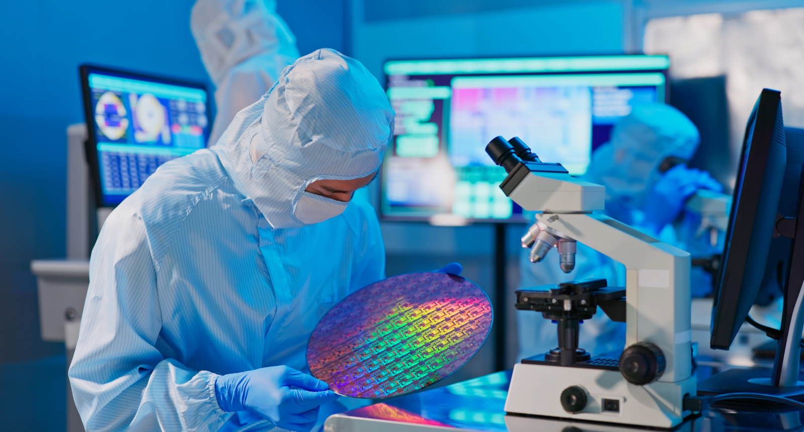 asian male technician in sterile coverall holds wafer that reflects many different colors with gloves and check it at semiconductor manufacturing plant